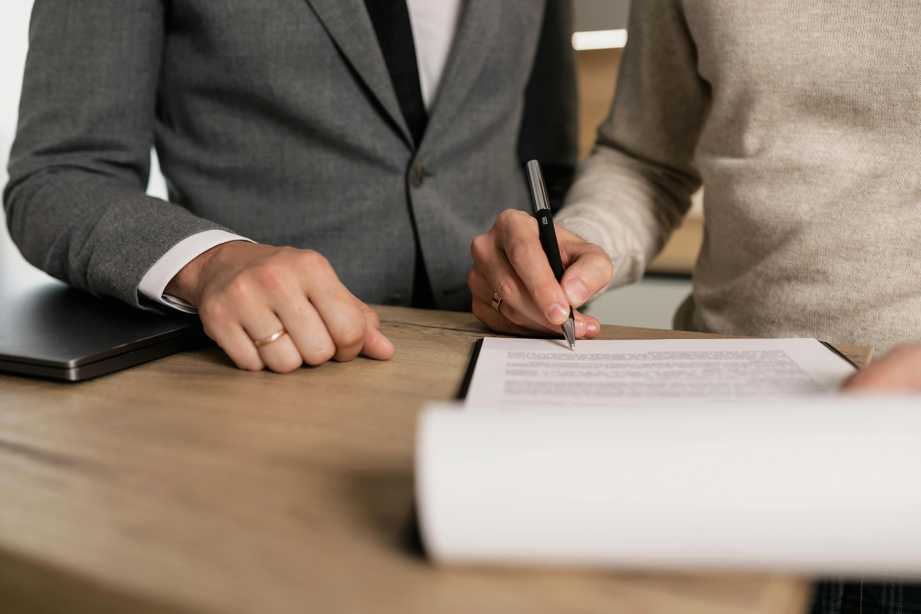 A close-up of two people's hands over a desk, one holding a pen and the other beside a notepad and documents.